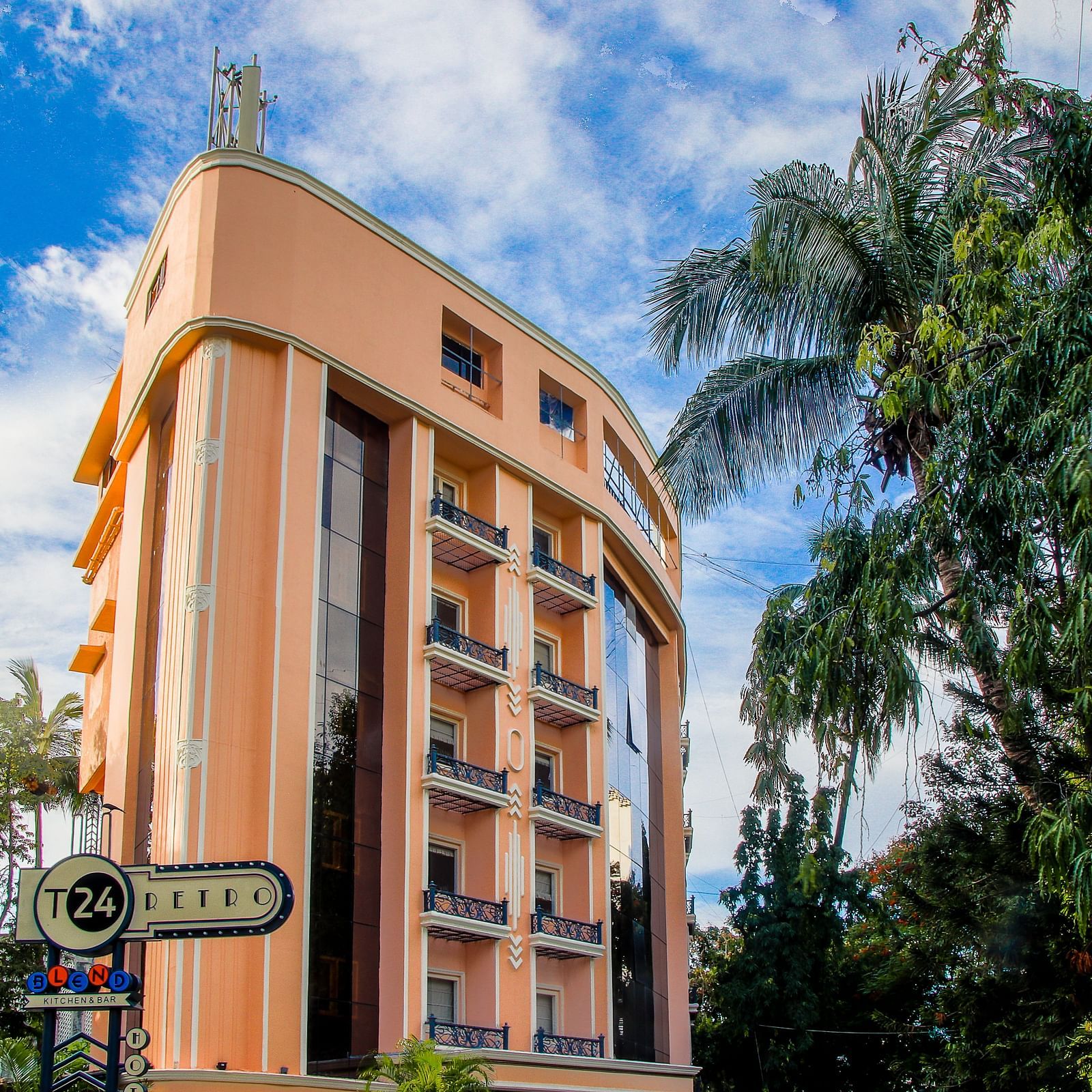 The exterior of the T24 Retro, a 4-star boutique hotel in Mumbai, India. The peach-colored building has a retro 70s style, with black balconies and large windows. Palm trees and other greenery line the street in front. 